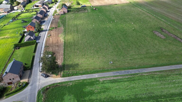 Vue aérienne d'un paysage rural à Ohey montrant un carrefour routier, des maisons adjacentes et des champs agricoles divisés à différents stades de culture.