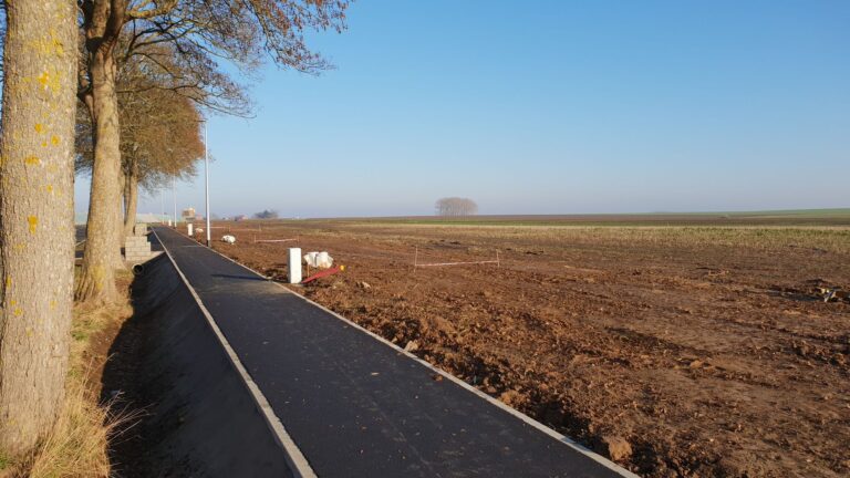 Une route nouvellement goudronnée à Taviers, à côté d'un champ fraîchement labouré avec des arbres nus sous un ciel bleu clair.