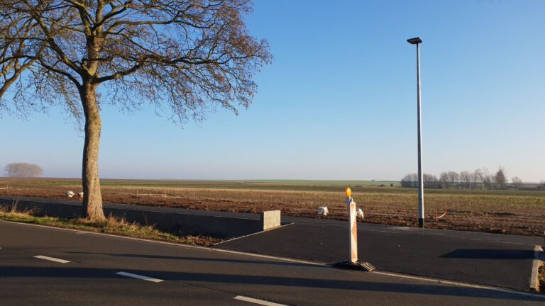 Vue sur une route rurale à Taviers, en Belgique, montrant une route nouvellement construite avec une piste cyclable, entourée de vastes champs et d'un ciel bleu clair.