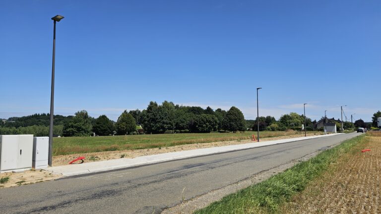 Un trottoir nouvellement pavé longe une route rurale avec des réverbères, des champs herbeux et des arbres sur un terrain de 12 ares sous un ciel bleu clair.