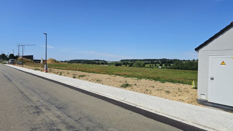 Une route pavée avec un trottoir longe un terrain dégagé sous un ciel bleu clair ; une boîte de services publics grise se trouve sur la droite, des grues de construction sont visibles au loin, et plusieurs maisons à quatre façades parsèment l'horizon.