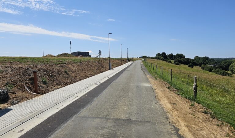 Une route pavée avec un trottoir sur le côté gauche traverse un terrain marécageux avec des champs herbeux et quelques bâtiments à quatre façades au loin sous un ciel bleu clair.