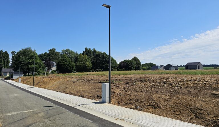 Un trottoir pavé longe une rue à côté d'un terrain dégagé avec un seul lampadaire ; des maisons avec 4 façades et des arbres sont visibles à l'arrière-plan sous un ciel bleu clair.