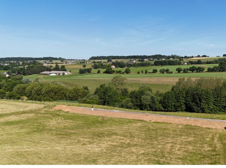 Une large vue d'un terrain rural avec des champs verts, des arbres épars et des maisons éloignées sous un ciel bleu clair, s'étendant sur 12 ares avec un charme pittoresque.