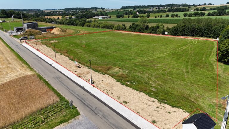 Vue aérienne d'un terrain de 12 ares à Hamois, délimité par un contour rouge, situé le long d'une route goudronnée et entouré de champs et d'arbres.