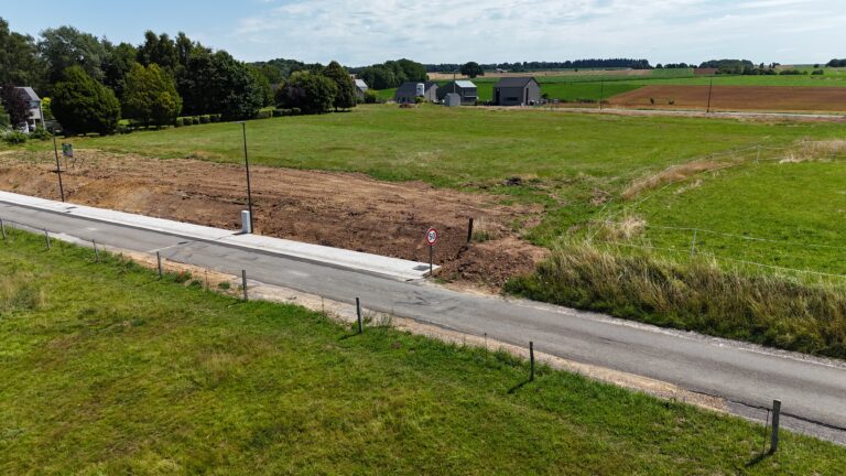 Un chemin rural longe un terrain en terre fraîchement défriché de 12 ares à Hamois, bordé de champs herbeux, de maisons éparses aux 4 façades, sous un ciel partiellement nuageux.